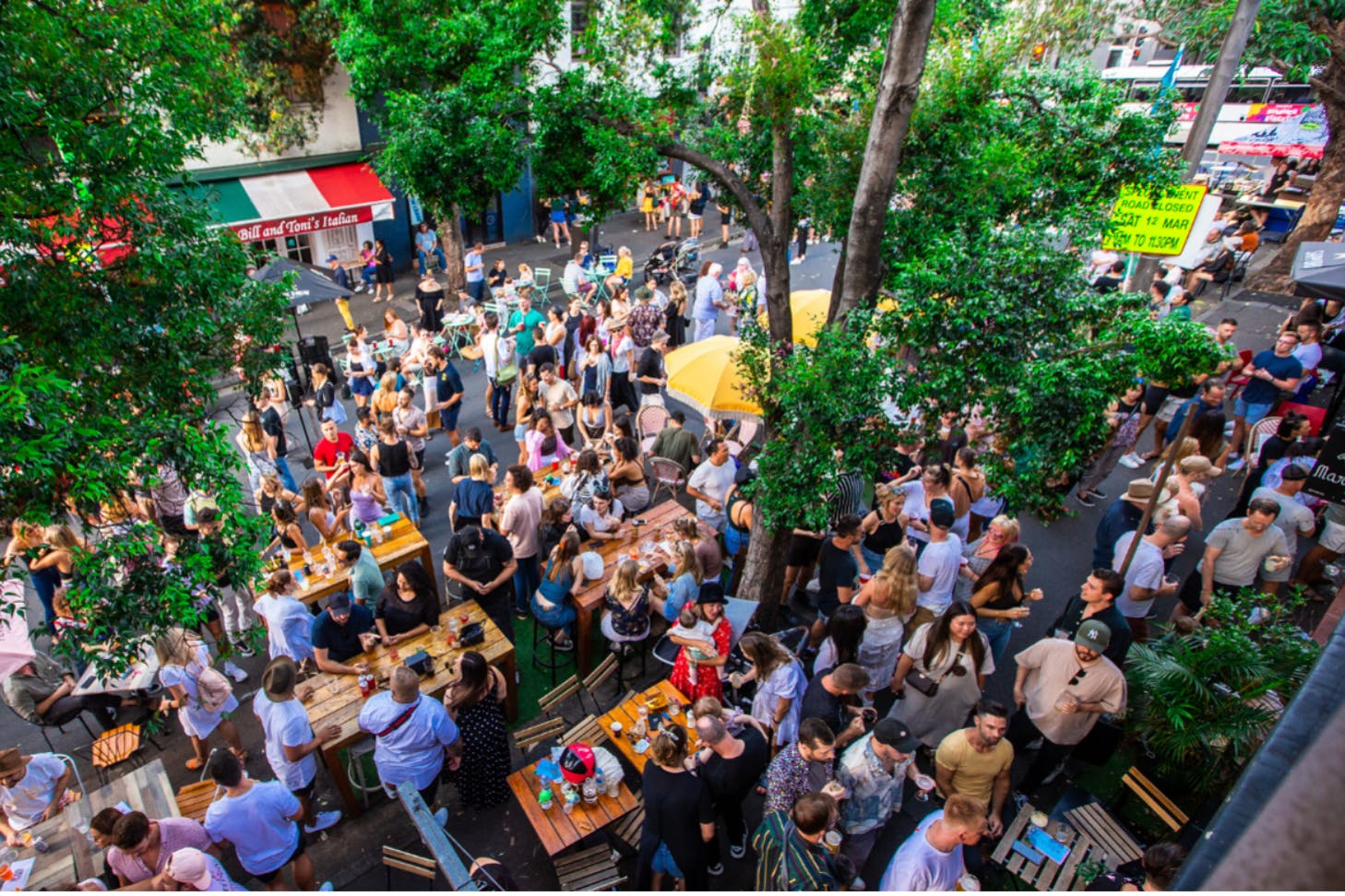 A wide shot from bird's eye view of a large crowd of people sitting outdoors at tables and walking around. People are chatting and socialising, the weather is nice and trees are around.